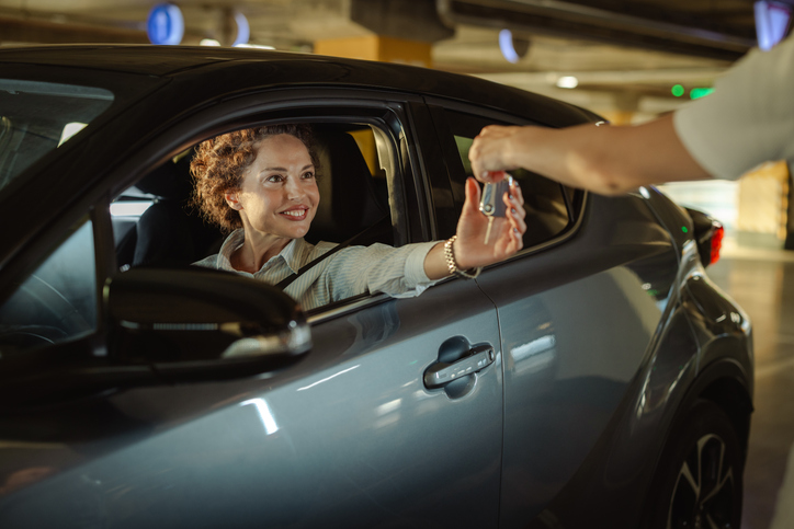 A happy individual receives car keys from another person while seated in a compact car, showcasing automotive transactions or rental services in a covered parking environmen